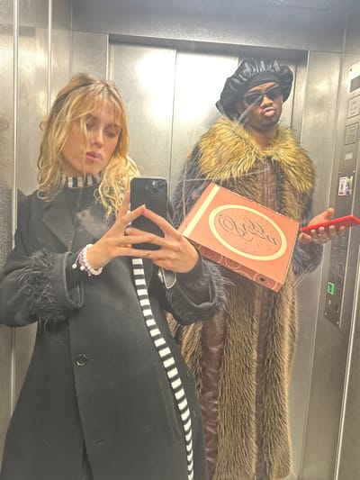 Two individuals are visible in a mirrored elevator setting. One person, positioned higher in the frame, wears a fur-trimmed jacket, black headwear, and sunglasses, and holds a red mobile phone and a rectangular box. The other person wears a black coat, takes a selfie and stands closer to the bottom of the frame. The elevator's interior is metallic, featuring control panels. The setting is suggested to be in Pantin, France.