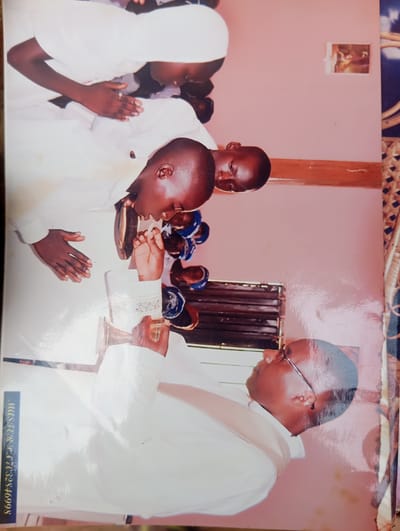 In Dutsen Alhaji, Nigeria, a man wearing glasses and a white robe is leaning forward. He is holding a golden object with his right hand. To his left, a young boy in a white robe and head covering is also leaning forward, appearing to be interacting with the man. The boy's left hand is extended. The background is a pink wall with a wooden shelf or ledge. To the left of the image, a blue strip with white text reads "MASTER R. T. 77632346998". The scene appears to be indoors, possibly a church or a formal gathering.