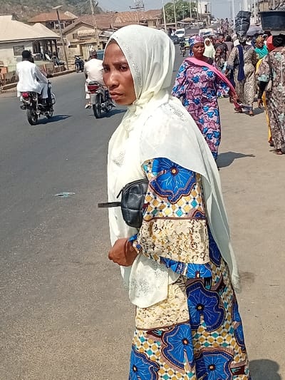 A woman wearing a white hijab and colorful dress stands on a street. Other women in similarly vibrant attire are nearby. Motorbikes and buildings line the background. The scene suggests a bustling outdoor environment, likely in a sunny location.