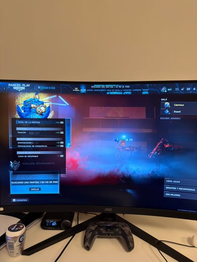 The image shows a person playing a video game, likely a first-person shooter, on a computer monitor. The game displayed is "Call of Duty: Warzone," indicated by the "WARZONE" text and the "RANKED PLAY" mode. The screen shows the post-game stats and matchmaking screen. The game appears to be in Spanish, with text like "BUSCANDO UNA PARTIDA" (Searching for a match) and "ELIMINACIONES" (Eliminations).
The user is in a room with a white wall and a black monitor. A black and gray camouflage controller is visible on the right side of the image, resting on a black stand. There are also some cables and a can of "Extra" chewing gum on a white surface. The lighting suggests it is indoors, possibly in the evening or a dimly lit room. The overall scene conveys a casual gaming session. The user's location is Frankfurt, Germany.