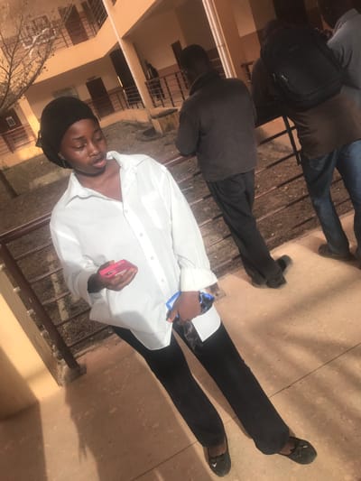 Three people are visible in this somewhat awkwardly angled image. One person stands in the foreground, wearing a white shirt, black pants, a black head covering and holding a pink phone in one hand and a blue phone in the other. Behind him, another person sits on a set of steps, wearing a dark jacket and pants. A third person sits on the steps to the top right and has on a brown jacket and blue jeans.