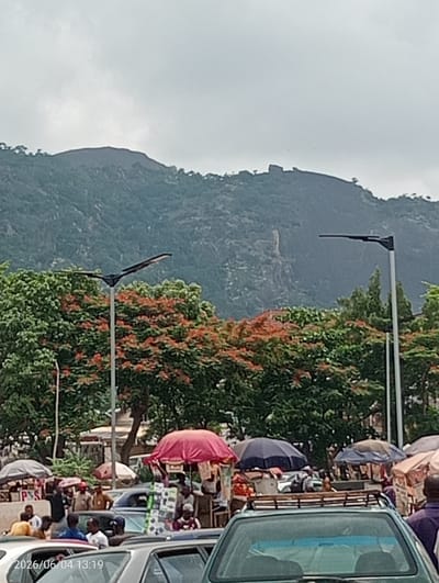 The image depicts a bustling street and market scene in Madalla, Nigeria, set against a backdrop of verdant mountains under an overcast sky.

In the foreground and mid-ground, numerous people are gathered amidst cars, suggesting a lively market or roadside commerce. Several cars are visible, including a silver sedan, a green vehicle with a roof rack, and others partially obscured. Market stalls, characterized by a variety of colorful umbrellas in shades of red, blue, brown, and white, are spread across the area, indicating places where vendors sell goods. Individuals are seen standing, walking, and interacting, with some appearing to engage in transactions.

Prominent in the mid-ground are several large trees, some of which feature striking clusters of orange-red blossoms, likely Flame Trees (Delonix regia), contrasting with the dominant green foliage. Tall streetlights with distinctive, flat, wide luminaires (possibly solar-powered) stand tall, extending above the tree line.

The background is dominated by large, rolling mountains or hills, heavily covered in green vegetation, particularly noticeable in the upper half of the frame. The sky is uniformly grey and cloudy, indicating overcast weather conditions.

A digital timestamp "2026/06/04 13:19" is visible in the lower-left corner, dating the image to early afternoon. A partial white sign displaying "P S" can be seen near one of the market stalls. The overall scene conveys a sense of vibrant community activity integrated with the natural landscape.