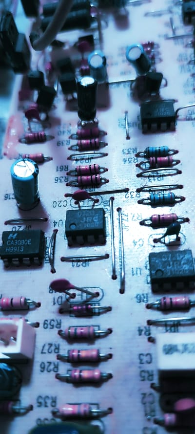 This close-up image captures a detailed view of a pink-hued electronic circuit board, likely part of an electronic device, in Remedios de Escalada, Argentina. The scene is static, showcasing an arrangement of various electronic components rather than any active event.
The board is densely populated with through-hole components, seen from a slightly elevated angle with a shallow depth of field, bringing some elements into sharp focus while others are softly blurred. Predominant subjects include numerous axial-leaded resistors, distinguishable by their colorful bands (blue, pink, brown) and labels such as R35, R26, R27, R59, R6, R4, R32, R33, R34, R36, R39, R40, and R61. Several electrolytic capacitors with cylindrical bodies (some with blue caps) are visible, along with labels like C2, C3, C4, C16, and C20.
Integrated circuits (ICs) in DIP (Dual In-line Package) form are scattered across the board. One IC prominently displays "CA3080E" and "H9913", identifying it as a CA3080 Operational Transconductance Amplifier. Another IC is marked "JRC 0720" and "97020", possibly a JRC4558 operational amplifier. Diodes are also present, one labeled D1. Several jumper wires are visible, marked JP12, JP19, JP20, JP21, and JP22, indicating configurable connections on the board.
The image is characterized by a strong cool blue tint, suggesting artificial lighting conditions or a specific filter. The overall impression is one of intricate electronic engineering, possibly under inspection or repair. No people are visible. Other minor text includes "10" and "CE".