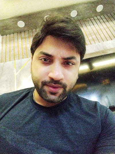 A medium shot captures a man with dark hair and a well-groomed beard. He wears a dark t-shirt and a gold chain is visible at his neckline. The background features a textured wall with vertical gold lines and recessed lighting. A blurred object, likely a television, is to his right. The lighting in the image appears to be artificial, casting shadows on his face. He gazes directly at the camera.