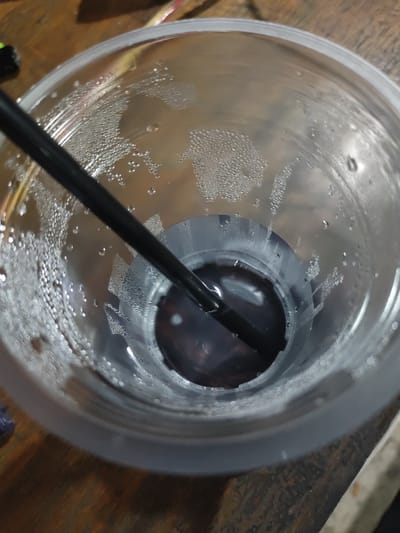 A close-up, overhead shot shows a clear plastic cup with condensation droplets on the interior walls. A black straw extends into the dark liquid at the bottom of the cup. The cup sits on a rustic wooden surface. The image appears to have been taken indoors, possibly in a home or cafe, but no specific location in Keudematangkuli, Indonesia can be identified from the visual evidence alone.