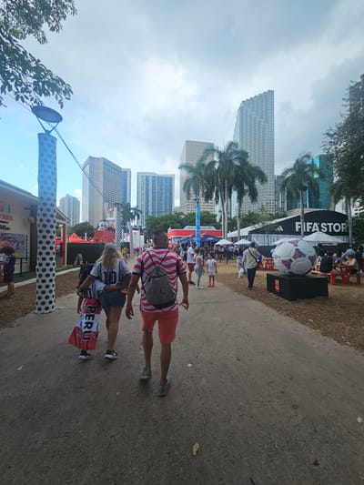 The image shows a lively outdoor event in a city. In the foreground, a family walks away from the camera. The father wears a red and white striped shirt and red shorts. The mother wears a white jersey with "MESSI 10" on the back and denim shorts. A child, likely their daughter, is between them, carrying a Peruvian flag.
The background reveals a cityscape with tall skyscrapers, including some with distinctive geometric designs. Palm trees are visible, suggesting a warm climate. The event appears to be football-themed, with a large replica of a soccer ball prominently displayed near a FIFA store. Other tents and booths, possibly selling merchandise or food, are set up around the area. People are milling about, creating a bustling atmosphere. The ground is a paved path, with some dry leaves scattered around, indicating it could be a park or public space. Based on the architecture and the presence of FIFA-related activities, the location is likely a public space in downtown Miami, Florida, United States, hosting a FIFA event.