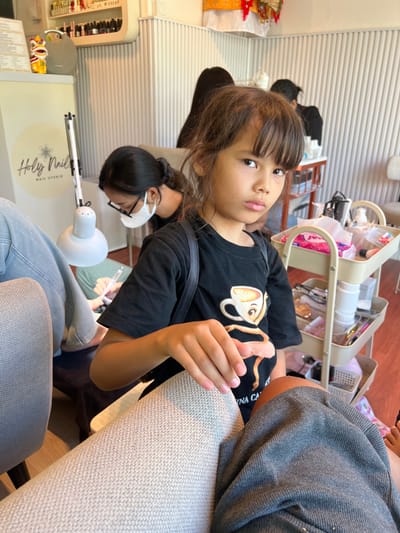 A young girl with dark hair and bangs, wearing a black t-shirt with a coffee cup graphic, sits in the foreground of the image. She is looking directly at the camera with a neutral expression. Her right hand rests on a textured light-colored fabric, likely a chair or cushion. In the background, a woman wearing a black t-shirt and glasses is seen working on someone's feet. She is wearing a white mask and holding a tool, suggesting a nail salon environment. A nail lamp is positioned over the feet. Other individuals and a multi-tiered trolley with various beauty products are also visible in the background, indicating a busy salon setting. The overall environment appears to be a well-lit indoor space, possibly a nail studio in Gianyar, Indonesia, given the "Holy Nail Studio" logo visible on signage.