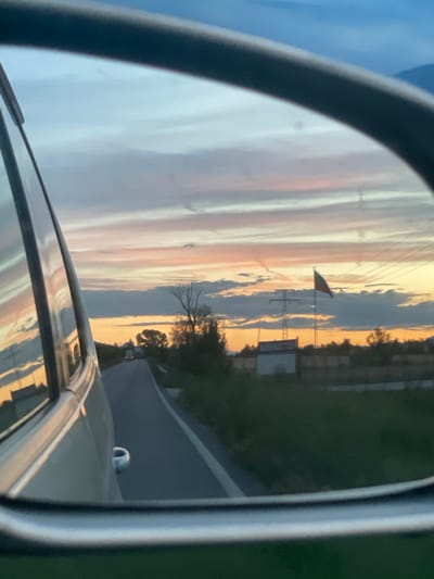 The image is a rear-view mirror reflection of a road at sunset. The sky displays a gradient of colors, from pale blue at the top to orange and pink near the horizon, with wispy clouds. A road stretches into the distance, flanked by green fields and trees. In the mid-ground, a power line tower is visible, and a flag with red, green, and yellow colors flies on a pole. The reflection also shows the side of a car, its metallic surface catching the colors of the sky. The scene appears to be in a rural area, possibly on the outskirts of Dabnitsa, Bulgaria, given the time of day and the agricultural landscape. No people are directly visible in the reflection.