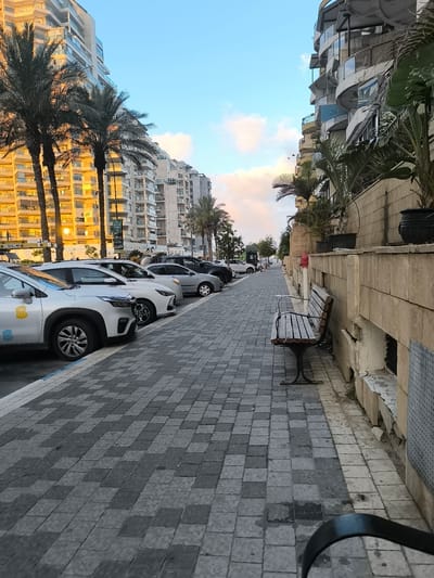 The image depicts a street scene in Ashkelon, Israel, with a sidewalk paved with dark and light gray interlocking square tiles. Palm trees line the left side of the street, with their fronds extending upwards. Behind the palm trees, a modern, multi-story apartment building with balconies is visible, its facade illuminated by warm light. Several white and silver cars are parked along the curb on the left side of the street.
To the right of the sidewalk, there is a low beige stone wall with planters containing green foliage. A dark wooden bench with black metal legs is positioned on the sidewalk in front of the wall. Further to the right, the scene is dominated by another multi-story building with curved balconies, partially obscured by palm trees and other lush greenery. The sky above is a light blue with scattered clouds tinged with pink and orange from what appears to be either sunrise or sunset. A red fire hydrant is visible near the bench.