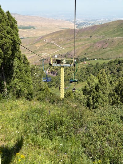 The image depicts a ski lift with several chairs on a sunny day in a mountainous landscape near Kashka Suu, Kyrgyzstan. The ski lift is yellow and is supported by a large yellow pylon. Several chairs, colored blue and green, are attached to the cable. Some chairs are occupied by people. In the foreground, there is lush green grass and vegetation. Behind the ski lift, rolling hills with winding roads and sparse trees are visible. In the far distance, a city or town can be seen with buildings and a hazy sky. The overall scene suggests a recreational area in a natural setting.