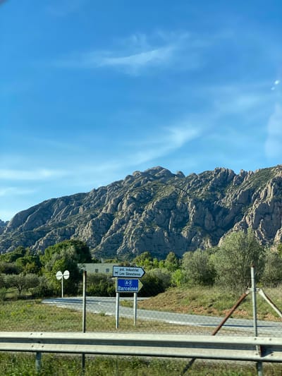 A roadside scene in Esparreguera, Spain. A rocky mountain occupies the left half of the frame, contrasted by a clear blue sky. On the right, a highway with guardrails is visible, accompanied by a sign indicating directions to "Pol. Industrial Les Ginestres" and "A-2 Barcelona." The landscape is a mix of rugged terrain and greenery, emphasizing the contrast between the natural environment and the built infrastructure.