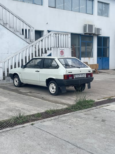A white, hatchback car is parked on a paved area next to a building. The building is white with blue-trimmed windows and metal staircases on the exterior. Two air conditioning units are mounted on the side of the building above a window. There is a "NO PARKING ZABRANJENO" sign on the building. The car has a Hungarian license plate that reads "AGN-894". The scene appears to be an outdoor industrial or commercial setting. The ground is concrete with a metal grate and some grass growing alongside it. The image is taken from a slightly elevated perspective, looking down at the car and the building. The lighting suggests it is daytime, possibly overcast, as there are no strong shadows.