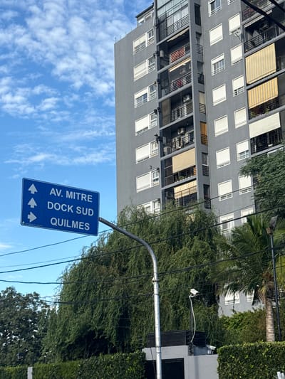 An urban scene captured in Avellaneda, Argentina, on a bright, sunny day. The dominant feature is a tall, multi-story building on the left side of the frame. The building is painted grey and features numerous balconies with dark railings, some equipped with air conditioning units. Many windows are fitted with white roller shutters, and several balconies have extended striped yellow and white awnings.
To the right of the building, a lush canopy of green trees and bushes extends, partially obscuring other structures and contributing to a green, treelined street atmosphere. A prominent blue street sign with white text and directional arrows hangs from a metal pole in the foreground on the right. Electrical wires are visible crisscrossing through the upper right portion of the frame, passing through the tree canopy.
The sky, visible in the lower part of the image, is a vibrant blue with scattered white, wispy clouds, indicating clear weather. No people or specific activities are visible.
Visible text on the sign reads:
AV. MITRE
DOCK SUD
QUILMES
Below these names are three white arrows, all pointing left, indicating directions to these locations.