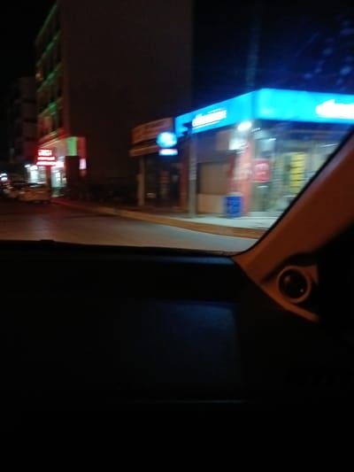 The image captures a night scene from inside a vehicle looking out at a street in Bahşılı Belediyesi, Turkey. A building with bright green and red neon lights is visible on the left, possibly a shop or restaurant. Across the street, a building with a prominent bright blue illuminated sign is present, which appears to be a service station or a similar establishment. The street itself is paved and appears to be moderately lit by the surrounding businesses. A yellow car is parked on the street. The interior of the vehicle is dark, with a visible speaker grille and a portion of the dashboard in the foreground. The overall impression is of a lively commercial area at night.