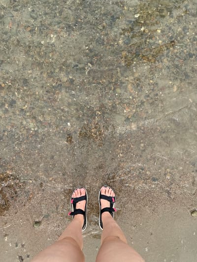 The image shows a person's feet standing at the edge of a body of water. The water is clear and shallow, revealing a rocky, sandy bottom. The person is wearing black sandals with pink accents. The sand on the shore is light brown and wet, with small pebbles scattered on it. The overall scene depicts a natural outdoor environment, likely a beach or the edge of a lake or river. There is no specific indication of the location in Tasheba, Russia, as the image lacks any distinguishable landmarks or contextual clues related to that city.