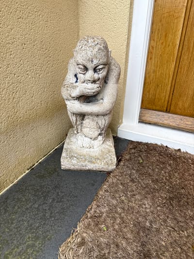 A stone gargoyle sits next to a doorway in Longden, United Kingdom. The gargoyle, weathered and showing signs of age with dark stains, is positioned on a textured, tan-colored wall adjacent to a white door frame and a light brown wooden door. A brown, rough-textured doormat is also visible. The photograph is taken from a close, elevated angle, revealing details of the surfaces and textures of the objects.