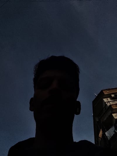 The image presents a low-angle shot, featuring a silhouetted individual in the foreground against a dark blue sky, located in Al-Qanawat Municipality, Syria. The subject, a person with discernible head and shoulders, is positioned centrally, facing slightly upwards, their features obscured by strong backlighting. To the right, a multi-story residential building with light-colored facades and dark-framed windows is partially visible. Laundry is hanging on a line from a lower balcony of this building. Several small, white satellite dishes are affixed to the upper sections of the structure. The sky is clear, with a deep blue hue indicative of twilight conditions.