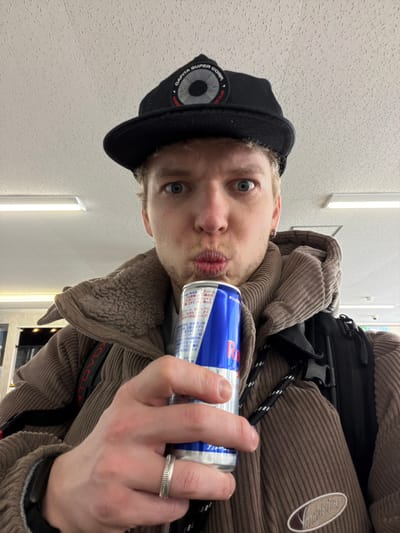 A fair-skinned man with light hair and blue eyes wears a black baseball cap and brown corduroy jacket. He holds a blue and silver Red Bull can near his pursed lips, gazing directly at the camera. A black backpack is slung over his shoulder. He wears a textured silver ring on his finger. The background features a white wall with rectangular light fixtures.