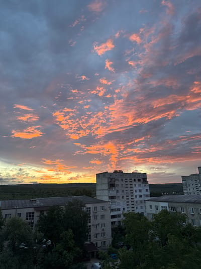 The image depicts an urban landscape in Strășeni, Moldova, dominated by a dramatic sunset. The sky is filled with vibrant orange and pink clouds, interspersed with darker gray clouds, creating a striking contrast. Below the sky, a row of multi-story apartment buildings, typical of Eastern European urban architecture, are visible. These buildings are constructed from concrete and brick, with numerous windows and balconies. Trees and greenery are scattered between and around the buildings, some appearing dense and lush. In the distance, rolling hills covered in vegetation form the horizon line. The overall scene conveys a sense of twilight in a residential area, with the natural beauty of the sunset contrasting with the man-made structures. There are no visible people or ongoing actions.