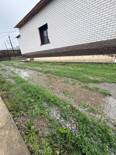 The image depicts an outdoor scene near a white brick house with a dark brown roof and trim. The house has a single dark-framed window visible. The ground in front of the house is covered in patchy green grass and gravel, with several areas showing standing water and what appears to be light rainfall. A wooden fence is visible to the left of the house, and power lines are strung overhead. The overall impression is of a somewhat unkempt yard in wet weather, likely in a rural or suburban setting within Солонешное, Russia. There are no people or animals present in the image.