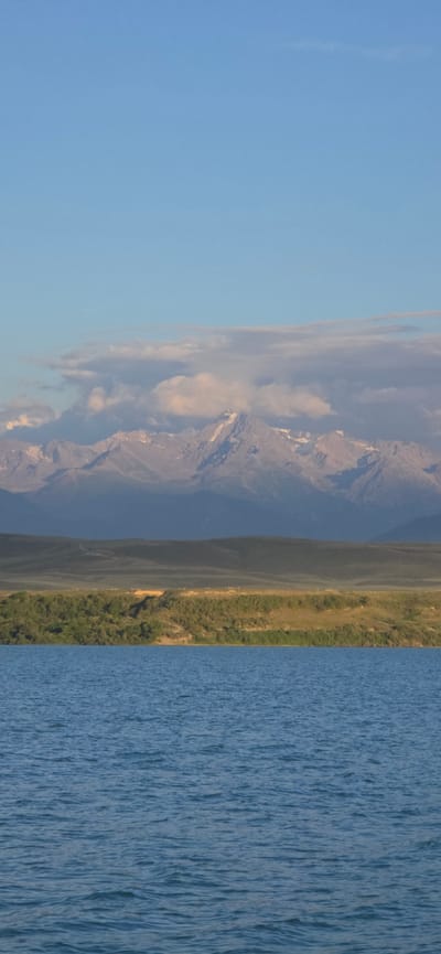 A serene landscape captures the vast expanse of a body of water in the foreground, its surface rippling gently under a clear blue sky. Beyond the water, a strip of verdant green foliage lines the shore, giving way to rolling hills that recede into the distance. The horizon is dominated by a majestic mountain range, its snow-capped peaks catching the soft light of the sun, with a scattering of clouds drifting above. The scene evokes a sense of tranquility and natural beauty, typical of the mountainous regions surrounding Lake Issyk-Kul near the city of Cholpon-Ata, in the Issyk-Kul Region of Kyrgyzstan.