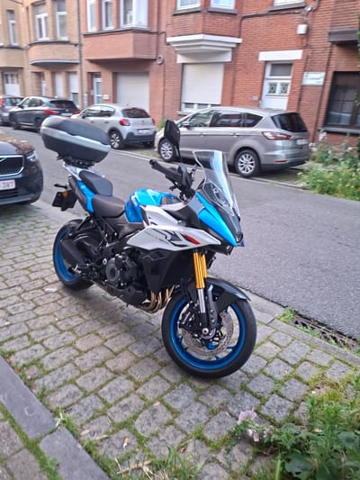 A blue and white Suzuki motorcycle is parked on a cobblestone sidewalk next to a street. The motorcycle is the primary subject, with its front wheel and handlebars visible, as well as its blue rim and gold suspension. A silver car is parked behind the motorcycle, and a brick building is to the left of the scene. The ground is a mix of cobblestones and grass, with some greenery visible. The time of day appears to be late afternoon or early evening, given the lighting. There is no visible text in the image. The scene appears to be a quiet street in a residential area, likely in Evere, Belgium.