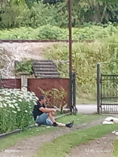 A man is sitting on the ground in front of a low wall, with a patch of white flowers to his left. He is wearing a dark t-shirt and blue shorts, and is looking to his right. Behind him, a brick wall is visible, with stairs leading up to it. A metal pole stands near the man, and a black metal gate is in the background. The scene appears to be outdoors, with lush green foliage in the background. The timestamp "28.06.2026 20:03" is visible in the lower right corner of the image. The location is indicated as Pshap, Abkhazia.