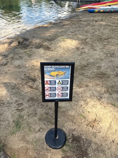 The image shows a sandy beach area next to a body of water. A sign is prominently displayed in the center of the frame, mounted on a black pole. The sign has a black frame and features a graphic of a yellow car driving on water, alongside text written in Polish. The text appears to be safety regulations related to water activities. The sign is positioned on the sand, with a few sparse green blades of grass growing in the foreground. In the background, the water's surface is visible, reflecting the sky. Further in the background, on the water's edge, several colorful paddleboards or kayaks are stacked. The overall environment suggests an outdoor recreational area, possibly a lake or riverfront.