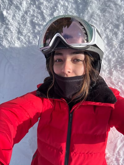 A person wearing a red snowsuit, ski goggles, and a black neck gaiter lies in the snow. The goggles reflect trees and sky. The person looks directly at the camera. The zipper of the snowsuit is partially open. The snow is bright and white.