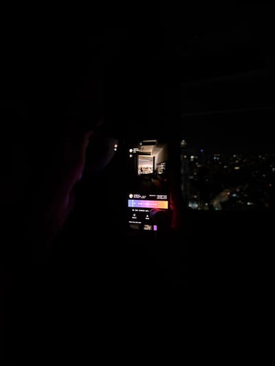 The image captures a dimly lit scene at night, taken indoors from what appears to be a high vantage point overlooking an urban area. The primary focus is a smartphone held in a person's hands, which are subtly illuminated by the phone's glowing screen. Beyond the hands and phone, the upper part of the frame reveals out-of-focus city lights, indicating a view through a window at night from Or Yehuda, Israel.

The smartphone screen displays a vertically oriented application interface. On the left side of the screen, a smaller, brighter image or video plays, depicting an indoor room with a prominent light source, possibly another screen or a brightly lit piece of art. The right side of the phone screen contains various interactive elements and text.

Visible text includes: "praceful, contemplative", "Introspective", "Sulpe/sligh to state", "No one staked yet,", "Share", "More from this u", and "Save". Icons for a down arrow, volume control, and potentially a 'like' or 'engage' button are also present. The screen's interface features a gradient of colors, including purple, orange, and pink hues. The overall impression is one of nocturnal digital engagement, with the phone's screen acting as the central light source in an otherwise dark environment.