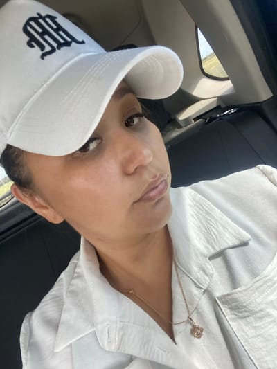A woman wearing a white baseball cap with a black monogram and a white collared shirt is seen from the chest up, looking at the camera. She has dark hair pulled back, brown eyes, and a neutral expression. A delicate gold necklace with a floral pendant rests on her shirt. The background shows the interior of a car, with a glimpse of a rural landscape through a window. The image appears to be taken inside a vehicle, possibly in motion, in Akyar, Russia.