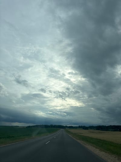 The image depicts a highway stretching into the distance under a dramatic sky filled with heavy, dark clouds. The road is asphalt with a white line marking the center. On the left side of the road, there is a vibrant green field, likely agricultural. On the right side, a field of dry, golden crop, possibly harvested wheat or hay, borders the road. A line of trees forms a dark silhouette against the sky in the far distance, marking the edge of the landscape. The overall scene suggests overcast weather, possibly leading up to or following a rain event. The location is identified as the Syarazhski Village Council, Belarus. There are no people or vehicles visible in the image.