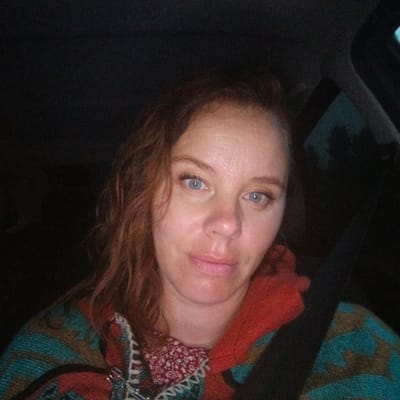 The image is a selfie taken by a woman in a car. She has fair skin, blue eyes, and reddish-brown wavy hair. She is wearing a colorful patterned poncho over a red floral shirt. The background is dark and appears to be the interior of a car, possibly at night, with some hint of the outdoors visible through the window. The overall lighting is dim. There is no indication of the specific location within the city of Кварса, Russia.
