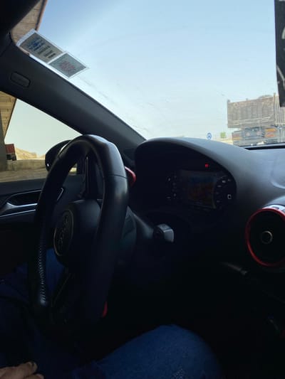 The image shows the interior of a car, likely an Audi, from the driver's perspective. The steering wheel, dashboard with a navigation screen, and part of the car's interior are visible. A sticker is affixed to the windshield. Outside the car, a large truck and some buildings are visible. The scene appears to be in Boughezoul, Algeria, as indicated by the sticker on the windshield which bears Arabic text and possibly registration details. The overall lighting suggests daylight.