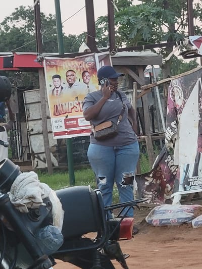 The image captures a scene likely in Suleja, Nigeria, given the "TANGOTE" (Dangote) cement bag. An individual wearing a cap and a sling bag is on a phone call, standing near a poster titled "LAMPST" featuring three men's faces, with event dates 13th and 14th at 4:30 PM and the words "PRAYER," "SOT," and "AM." There's also another sign with "PUTER NETWORK WORD" visible above other banners. A motorcycle is prominently featured in the foreground.