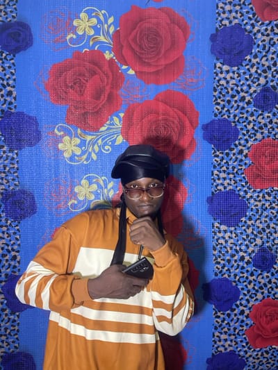 An individual is depicted lying on their back, looking directly at the camera. The person, appearing to be male with a dark complexion, is wearing a black cap, circular brown-framed glasses, and an orange long-sleeved shirt with vertical white stripes. One hand is positioned near their chest, holding a black cell phone, while a black strap (possibly from a bag or headphones) crosses their chest.
They are resting on a vibrant, patterned surface. This surface features a dominant blue background, adorned with large red roses and intricate golden floral or vine-like accents. Interspersed are horizontal sections with a different pattern: a leopard print motif against a blue background, accented with smaller, darker blue and red flower shapes. The texture of the surface suggests it could be a mat or a fabric with a distinct weave or print.
The lighting appears even, suggesting an indoor setting or a shaded outdoor area, within the context of Nkwelle, Nigeria. The person's expression is neutral, engaging directly with the viewer. There is no visible text in the image.
