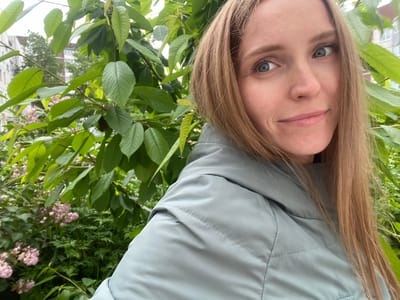 A woman with blonde hair and blue eyes, wearing a light gray jacket, poses for a selfie in front of lush green foliage. Behind her, glimpses of buildings and pink flowers can be seen. The scene appears to be outdoors in an urban environment.