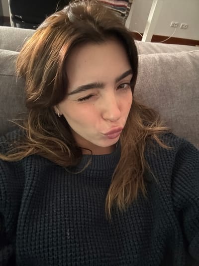 The scene depicts an indoor setting, located in Nogueira e Silva Escura, Portugal. A young adult female is visible from the upper chest upwards, positioned centrally in the frame. She has long, wavy brown hair with lighter highlights. Her left eye is closed in a wink, and her lips are pursed in a pout, directed towards the viewer. Her skin shows a slight flush on the cheeks. She is wearing a dark blue or dark grey knitted sweater with a prominent waffle or rib-knit texture. Small silver hoop earrings are visible in her ears. The subject is resting against a grey upholstered surface, likely a sofa or cushion, which occupies the background behind her. In the upper right portion of the background, a light-colored wall features two white electrical outlets and a horizontally running brown cable. A small section of multicolored striped fabric is partially visible in the upper left corner of the background.