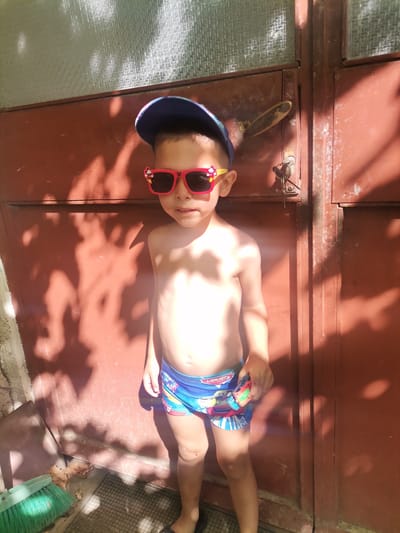 A young boy, likely Caucasian, stands shirtless and wearing swim trunks, a blue baseball cap, and pink sunglasses with cartoon characters on them. He is standing in front of a weathered, reddish-brown metal door with a visible door handle and lock mechanism. Sunlight casts strong shadows of foliage onto the door and the ground. To the left of the boy, a green broom and some dry leaves are visible on the ground. The scene appears to be outdoors, with a textured glass panel above the door. The location is identified as Obrochishte, Bulgaria.
