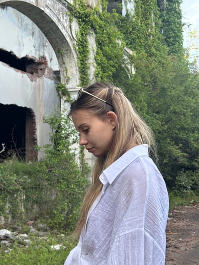 A young, blonde woman wearing a white button-up shirt stands facing downward in front of the ruins of a building. The building is primarily made of concrete and brick, with large openings and arches, and is partially covered in green vegetation. The location appears to be Tsanbaikwara, Abkhazia.