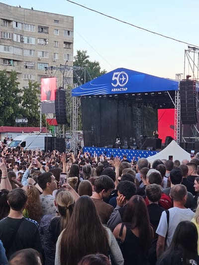 A large outdoor crowd of young people are gathered in front of a stage, holding up their phones and arms. The stage is set up with a large blue banner that reads "50 АВИАСТАР" with an airplane logo. There are large speakers on either side of the stage, and a big screen displaying an image of a man. Behind the stage, a tall apartment building is visible, with trees and a clear sky. In the foreground, a white van is parked, and a sign that reads "ЦВЕТЫ" is visible. The overall scene depicts an outdoor concert or festival event. Based on the banner "АВИАСТАР", this event is likely related to aviation and could be held in Ulyanovsk, Russia, as Ulyanovsk is a major center for the Russian aviation industry.