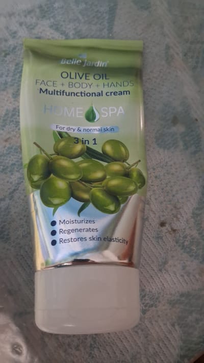 The media file displays a close-up, overhead view of a tube of "Belle Jardin OLIVE OIL FACE + BODY + HANDS Multifunctional cream" positioned on a textured surface. The tube is oriented horizontally with the cap on the right side. The product packaging is predominantly green and silver, featuring a graphic of green olives on a branch. Visible text on the tube includes "Belle Jardin," "OLIVE OIL," "FACE + BODY + HANDS," "Multifunctional cream," "HOME SPA," "For dry & normal skin," and "3 in 1." Additionally, three bullet points describe the cream's benefits: "Moisturizes," "Regenerates," and "Restores skin elasticity." The background surface appears to be a woven textile with a blue-green hue and a subtle pattern. There are no people or animals visible in the image, and the setting is ambiguous, suggesting an indoor environment given the focus on the product. The lighting is even, and there are no distinct time-of-day or weather cues. The context provided places this image in Beloci, Moldova.
