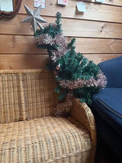 The image shows a small, decorated artificial Christmas tree on a wicker bench next to a dark blue chair. The tree is adorned with tinsel and a silver star on top. The background is a wooden wall with several notes attached. A single Christmas ornament rests on the wicker bench in the foreground. The scene appears to be indoors. There is no information in the image to suggest the location is in Oslo, Norway.
