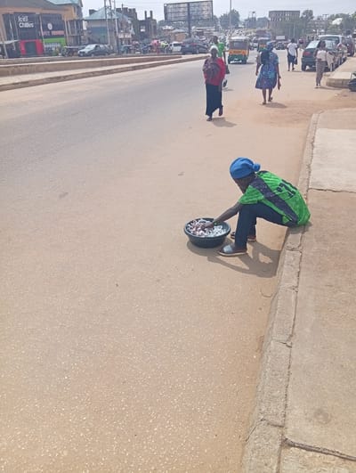 The image captures a street scene in Kabong, Nigeria, on a bright, sunny day. A young individual wearing a blue knit cap and a green and black patterned shirt is crouched at the edge of a concrete sidewalk, facing a black bowl filled with small, white objects. The individual appears to be a vendor, possibly selling the items in the bowl, which resemble small candies or snacks. They are wearing dark jeans and grey slide sandals. The sidewalk itself is worn and cracked in places.
In the background, the street is bustling with activity. Several vehicles, including a green car, a white van, and at least two auto-rickshaws (one yellow and one white), are visible on the asphalt road. Pedestrians are also present, with a woman in a blue and white patterned dress walking away from the camera, and another woman in a red top and black skirt walking towards it. Further down the street, more people and vehicles are present, along with buildings that appear to be shops and residences. The overall environment suggests a commercial or residential area within the city, with active daily life.