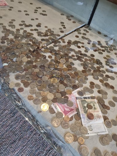 The image shows a collection of coins and banknotes on a clear surface, possibly a table or display case. There are hundreds of coins of various sizes and colors, scattered across the surface. Among the coins are two Bulgarian banknotes, one a 10 leva note featuring an image of Petar Beron, and the other a smaller denomination with a pink hue. Based on the banknotes, the location could be in Bulgaria. The image also shows a carpet next to the clear surface. Elena is not referenced in the image.