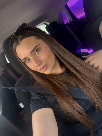 The image shows a young woman in the passenger seat of a car, facing the camera. She has long, dark brown hair that is styled straight and falls over her shoulders. She is wearing a black t-shirt and a seatbelt. Her gaze is directed towards the camera with a slight smile. In the background, the interior of the car is visible, with black seats and a purple light emanating from the ceiling or a window. The scene appears to be a selfie taken within the city of Razliv, Bulgaria.