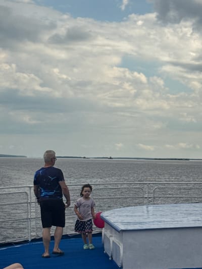 In Ikskoe Ust'e, Russia, on what appears to be a ferry or boat, a man and a young girl stand on a deck. The man, viewed from behind, wears a dark blue t-shirt with a graphic design and black shorts. He is facing away from the camera, looking out at a body of water. The girl, facing the camera, wears a light pink t-shirt and a dark patterned skirt. She stands to the right of the man, and a pink inflatable object is visible near her. They are positioned near a railing and a blue and white structure. The sky above is cloudy.