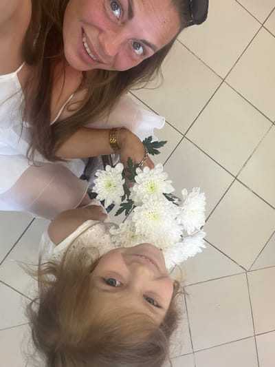 This is a vertical, low-angle, eye-level medium shot of two females posing for a selfie with a bouquet of white flowers. The female on the left is a young girl with light brown hair and blue eyes. She is wearing a white dress and has a sweet expression. She is holding a bouquet of white chrysanthemums. The female on the right is an adult woman with long brown hair, also wearing a white dress. She has a wide smile and is wearing a watch on her left wrist. The background consists of a tiled floor. The lighting is bright, suggesting it is daytime. The scene appears to be indoors. The overall mood is happy and affectionate.