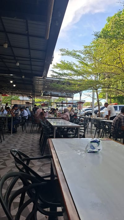 This is an outdoor scene at a restaurant in Keuniree, Indonesia, with tables and chairs set up under a covered roof. Several people are seated at tables, some eating, some conversing. A man in a white shirt is standing near a parked car, looking towards a table. Green trees are visible in the background, and the sky is partly cloudy. The flooring is tiled.