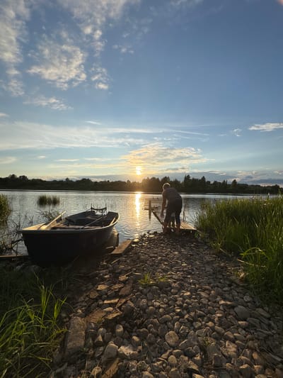 At sunset on a body of water in Sitno, Russia, a person is standing on a wooden pier, facing away from the camera. The person is wearing a striped shirt and dark pants. A small boat is moored to the pier, and tall grass grows along the water's edge. The sky is blue with scattered clouds, and the sun is setting on the horizon, casting a golden glow on the water. The shoreline is covered in stones and dirt.